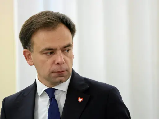 Minister finansów w granatowym garniturze i niebieskim krawacie, z przypinką w kształcie serca na klapie marynarki.