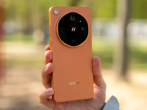 Pomarańczowy oppo flagowiec z charakterystycznym modułem aparatu w dłoni, na tle rozmytego, zielonego krajobrazu.