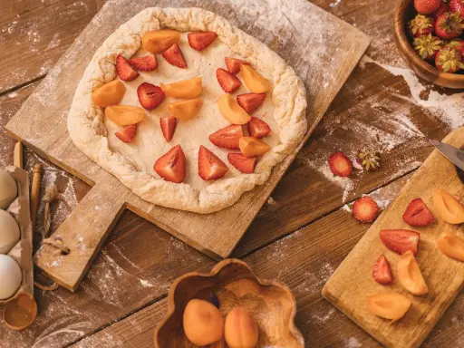 Préparation d'un crumble abricot et fraises sur une planche en bois, avec des œufs et des fruits frais à proximité.