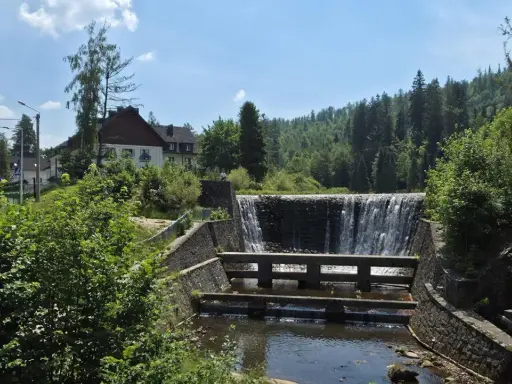 Kamienny stopień wodny na Jeziorze Czerniańskim, z wodą spływającą kaskadami. W tle zielone wzgórza i domy.