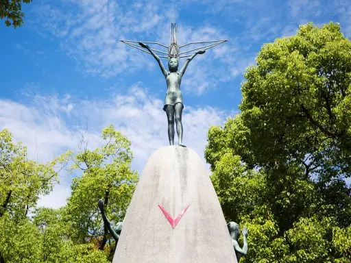 La bambina di Hiroshima, simbolo di pace, con le braccia tese verso il cielo, circondata dal verde degli alberi e dall'azzurro delle nuvole.