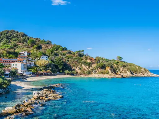 En pittoresk kustby på Elba, Italien, med vita hus som klättrar uppför en grönskande kulle. Klart turkost vatten sköljer mot en sandstrand.