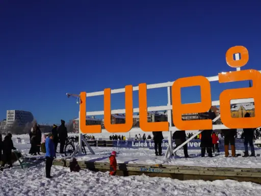 Stor orange skylt med texten "LULEÅ" i snöig miljö. Perfekt för luleå travel, en solig vinterdag.