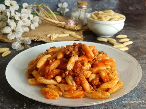 Un piatto invitante di malloreddus alla campidanese, conditi con un ricco ragù di carne. Sullo sfondo, fiori bianchi e un ciotola di pasta cruda.