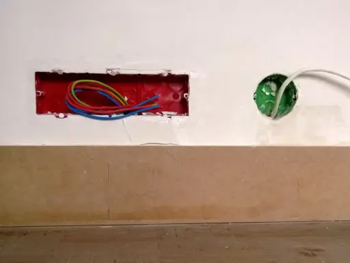 Boîtier rouge avec fils électriques colorés, prêt pour un interrupteur compteur électrique. Une boîte verte avec un câble blanc est visible à droite.
