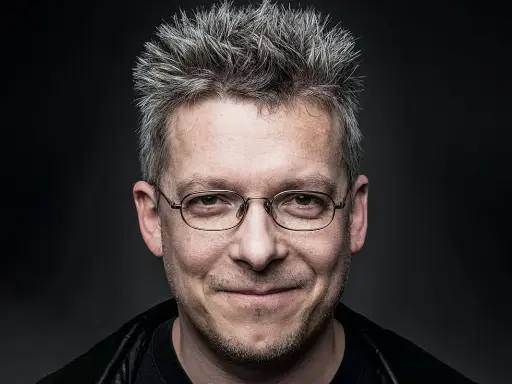 Uśmiechnięty mężczyzna w okularach, z siwymi, nastroszonymi włosami. Może to być jeden z bohaterów Felix Net i Nika, gotowy na przygodę.