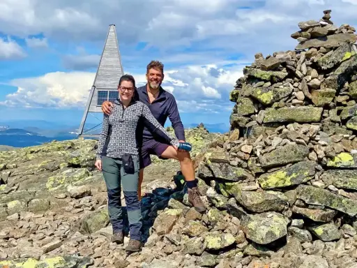 Två glada vandrare njuter av utsikten från en bergstopp under en vandring i Sverige. En stenröse och en modern träkonstruktion med solpaneler syns i bakgrunden.