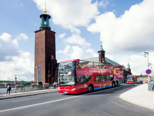 Röd dubbeldäckare med "Hop-on Hop-off Stockholm Buses" på väg förbi Stadshuset. Perfekt för stockholm turistinformation.