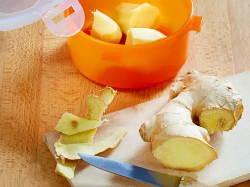 Frische Ingwerstücke werden geschält und in einem orangen Behälter aufbewahrt, um **Ingwer haltbar zu machen**. Ein Messer liegt daneben.