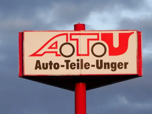 Schild mit rotem Schriftzug "A.T.U. Auto-Teile-Unger" vor bewölktem Himmel.
