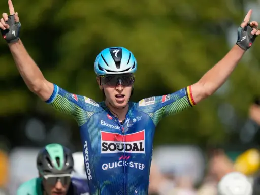 Tim Merlier jubelt mit erhobenen Armen und zeigt den Sieg. Er trägt ein blau-grünes Radtrikot mit Sponsorenlogos.