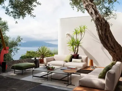 Arredare un terrazzo scoperto con divani chiari, tavolini bassi e un pouf verde, con vista mare e un ulivo secolare.