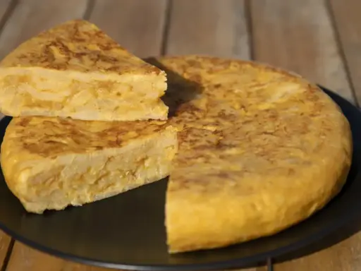 Una deliciosa tortilla de patatas, con una porción cortada y apilada, servida en un plato negro sobre una mesa de madera.