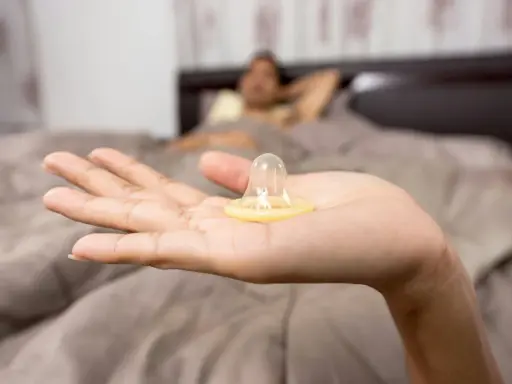 Mano sostiene un condón. En el fondo, una persona en la cama. La decisión de tener sexo oral sin condón es personal.