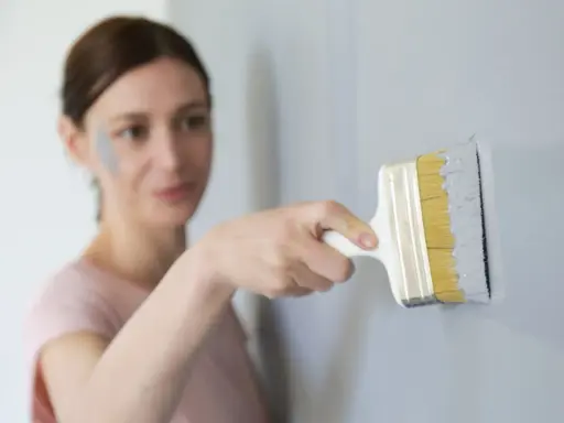 Une femme effectue une retouche peinture murale avec un pinceau gris. Une tache de peinture grise est visible sur sa joue.