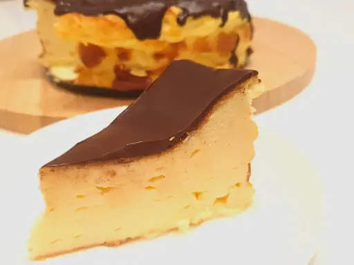 Kawałek puszystego keto sernika z mascarpone, polany czekoladą. Cały sernik w tle.