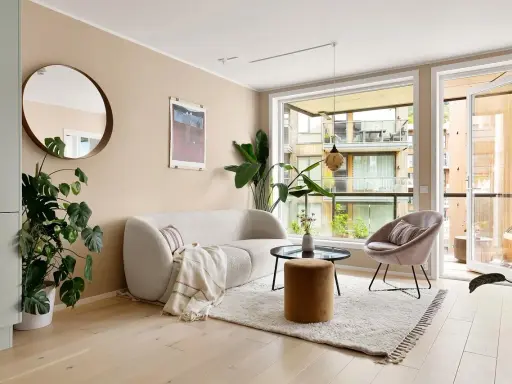 Jasne meble do salonu: puszysta sofa, okrągły stolik, aksamitny fotel i puf. Duże okno z widokiem na balkon.