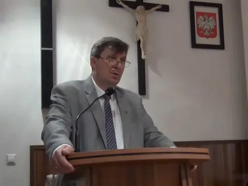 Mężczyzna w garniturze przemawia, za nim krzyż i godło Polski. Może mówi o cnotach kardynalnych.