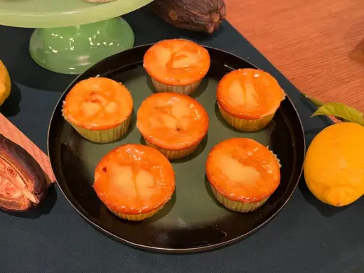 Sechs Zitronenmuffins mit glänzender Glasur auf einem Teller, daneben frische Zitronen und eine Kakaofrucht. Ein leckeres zitronenmuffins rezept!