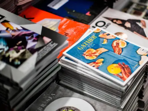 Stosy magazynów, jeden z nich z okładką "GATHER" i pomarańczami, gdzie każdy detal, od tekstury owoców po odbicia światła, podkreśla piękno koloru spotowego.
