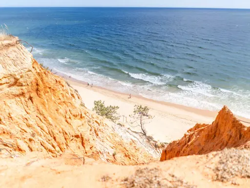 Orange cliffs overlook a sandy portugal strand algarve. Gentle waves lap the shore where a few people stroll.