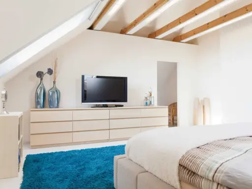 Schlafzimmer im Dachgeschoss, ideal zum Dachboden ausbauen. Helle Farben, Holzbalken und ein blauer Teppich schaffen Gemütlichkeit.
