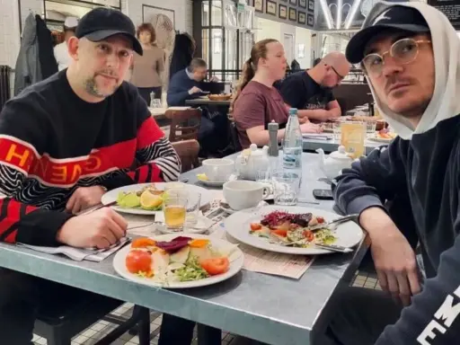 Dwóch mężczyzn, jeden w bluzie z napisem "Givenchy", drugi w bluzie "Supreme", siedzi przy stole w restauracji. Wyglądają jak bary raperzy, którzy wybrali się na wspólny posiłek.
