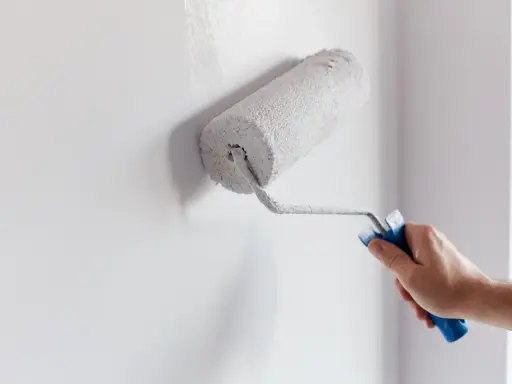Ręka maluje ścianę wałkiem, nakładając białą farbę. To prosty sposób, jak zamalować farbę ceramiczną, by odświeżyć wnętrze.