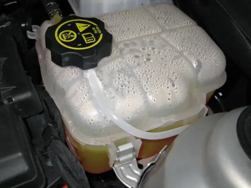 Depósito de expansión con líquido refrigerante y burbujas. La tapa negra tiene símbolos de advertencia.