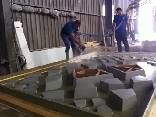 Dos operarios trabajan con material GRC en un taller, uno rocía con una manguera y el otro supervisa.