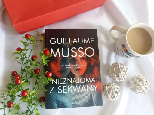 Książka "Nieznajoma z Sekwany" Guillaume'a Musso leży obok kubka kawy i czerwonych ozdobnych kulek.