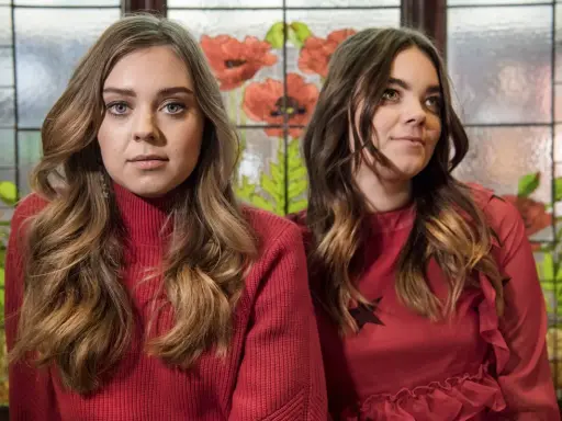 Två medlemmar av First Aid Kit poserar framför ett fönster med blommotiv.