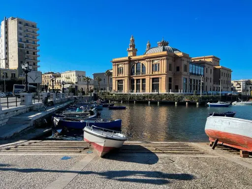 Barche ormeggiate nel porto di Bari, con il Varco della Vittoria e il Teatro Margherita sullo sfondo, sotto un cielo azzurro.