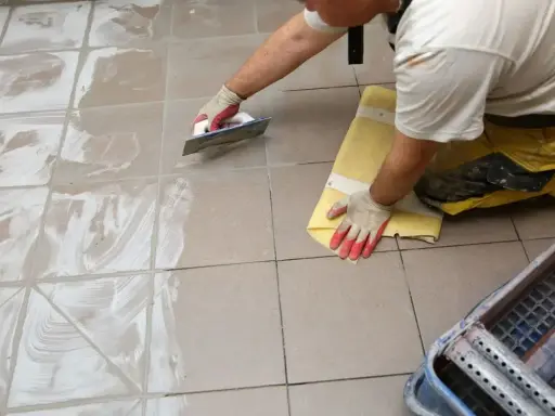 Pracownik w rękawiczkach nakłada fugę epoksydową na płytki podłogowe za pomocą kielni. Proces fugowania krok po kroku.