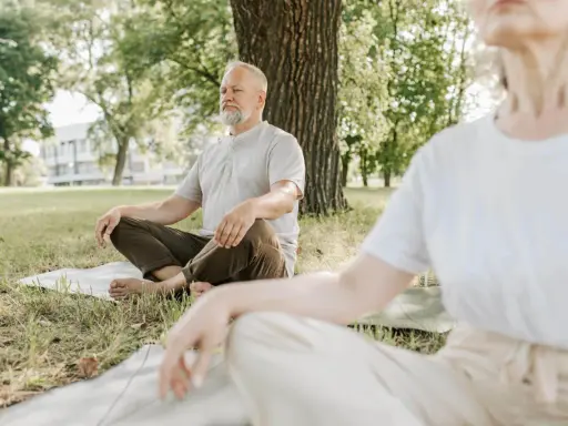 Två personer sitter i lotusställning på gräsmattan under ett träd. De utövar liggande meditation i en lugn parkmiljö.