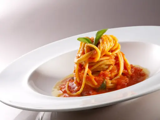 Un nido di spaghetti conditi con una salsa rossa vibrante di pomodorini freschi, guarnito con foglie di basilico verde brillante, servito in un piatto bianco elegante.