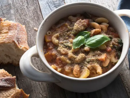 Una ciotola di pasta e fagioli in scatola, guarnita con basilico fresco e pane croccante su un tavolo di legno.