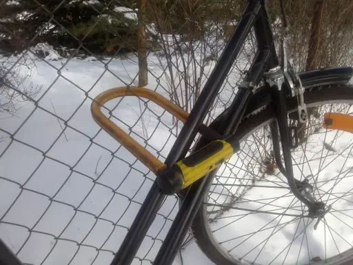 Żółta kłódka U-lock zabezpieczenie roweru przed kradzieżą, przypiętego do siatki w zimowy dzień.
