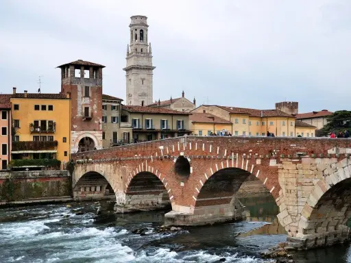 En vacker tegelbro korsar en flod i Verona, Italien. Färgglada hus och ett högt klocktorn syns i bakgrunden.