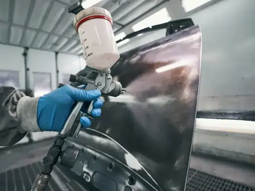 Ręka w niebieskiej rękawiczce trzyma pistolet lakierniczy, rozpylając farbę na element karoserii. Czas schnięcia szpachli na epoksyd jest kluczowy dla dalszych prac.