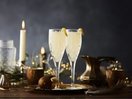 Två eleganta glas med en uppfriskande drink med gin och prosecco, dekorerade med citronskal. Stämningsfull belysning med ljus och ljusslingor skapar en festlig atmosfär.