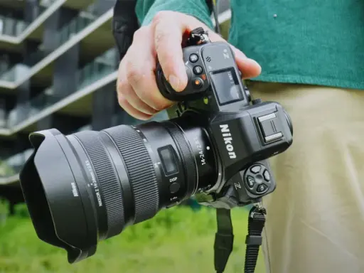 Dłoń trzyma aparat Nikon Z8 z obiektywem. W tle nowoczesny budynek.