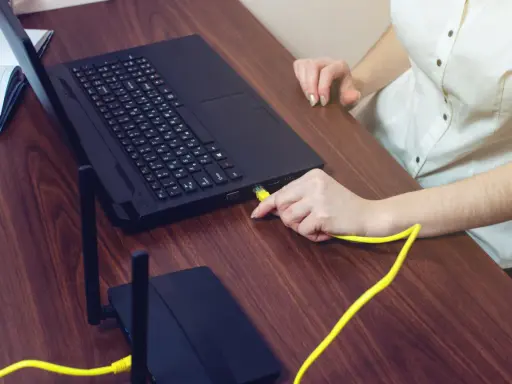 Ręka podłącza żółty kabel do laptopa, pokazując, czy można podłączyć monitor do laptopa. Obok leży router.