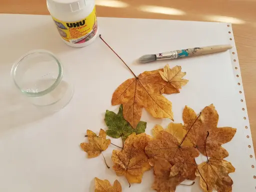 Préparation d'une décoration avec des feuilles d'arbre séchées, de la colle UHU, un pinceau et un petit récipient en verre.