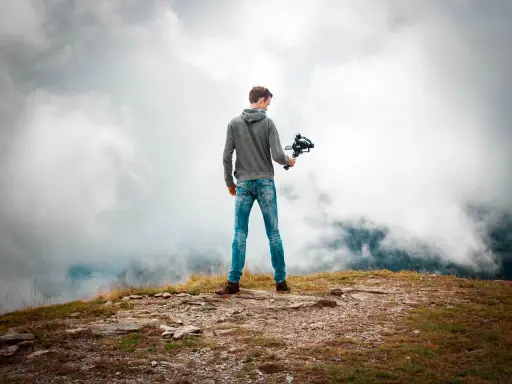 Joven en la cima de una montaña, sosteniendo un **gimbal** para estabilizar su cámara y capturar la vista entre las nubes.