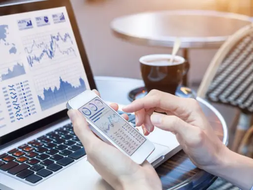 Manos revisando gráficos financieros en un teléfono móvil y portátil, gestionando el bbva net cash.