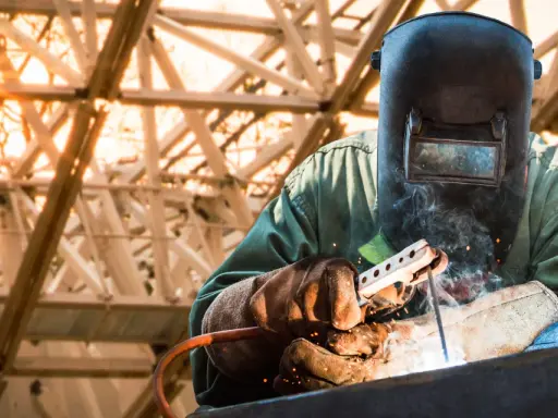 Soldador con máscara y guantes realiza soldadura con electrodo, creando chispas y humo bajo una estructura metálica iluminada por el sol.