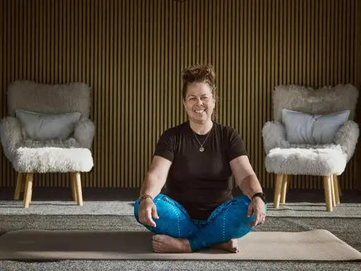 Kvinna i meditationsställning framför en vägg med träribbor. Två fluffiga fåtöljer står på var sin sida. Kanske hon funderar på hur hon ska dekorera hylla i sitt hem.