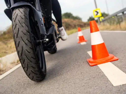 Motociclista practica maniobras entre conos naranjas. La rueda delantera de la moto, con su banda de rodadura, se ve de cerca.