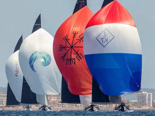 Segelboote der j class rasen über das Meer. Bunte Spinnaker mit Logos und Symbolen, im Hintergrund eine Küstenstadt.